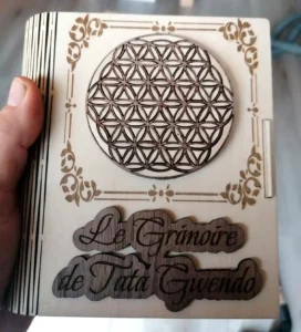 Boîte en bois en forme de livre, pour ranger un jeu d'oracle, avec motif fleur de vie en marqueterie deux essences de bois et texte découpé 'Le Grimoire de Tata Gwendo